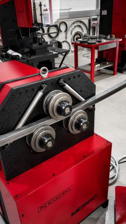 Máquina curvadora industrial roja con tres rodillos curvando un tubo metálico cuadrado en un taller.